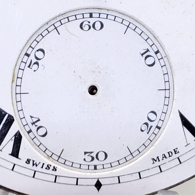 May include: Close-up of a white watch face with black numerals marking the hours and minutes. The watch face has a small hole in the center and the words "SWISS" and "MADE" are visible.
