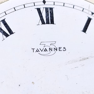 May include: Close-up of a watch face with a white background. Black Roman numerals mark the hours, with "XII" prominently displayed. The word "TAVANNES" is printed in black, beneath the numerals, inside a stylized emblem.