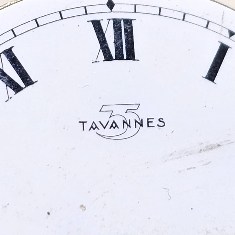 May include: Close-up of a watch face with a white background. Black Roman numerals mark the hours, with "XII" prominently displayed. The word "TAVANNES" is printed in black, beneath the numerals, inside a stylized emblem.