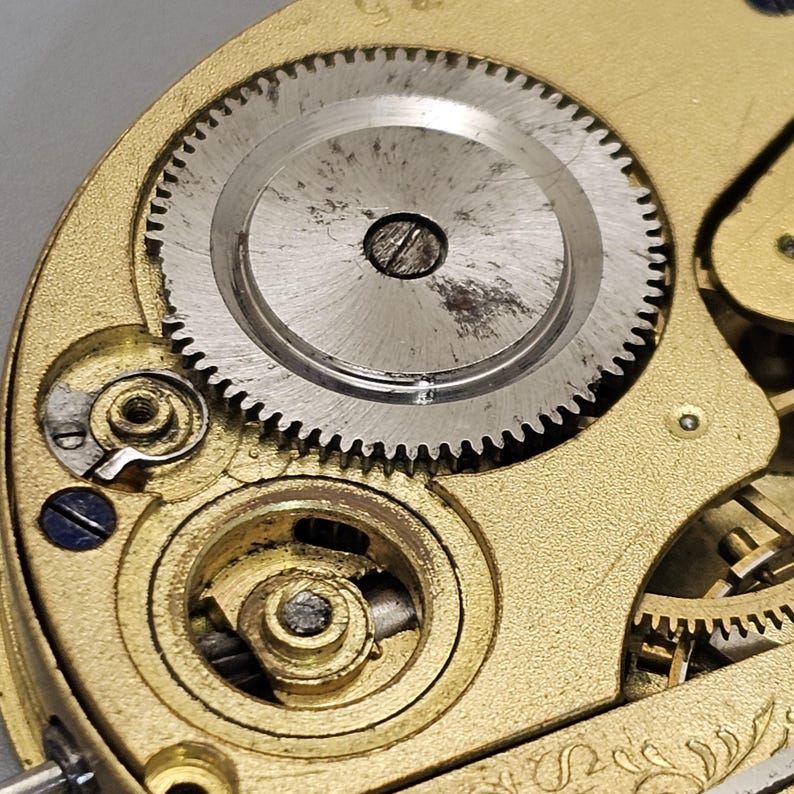 May include: Close-up of a gold-colored watch mechanism with a silver gear and a small black screw in the center.
