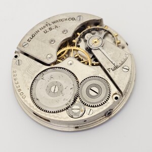 以下が含まれることがあります： Elgin National Watch Co. U.S.A. の時計ムーブメントのクローズアップ。 銀色のメカニズムは、複雑な歯車、ネジ、番号2297600を表示します。 このヴィンテージの時計ムーブメントは、時計の内部コンポーネントを明らかにしています。