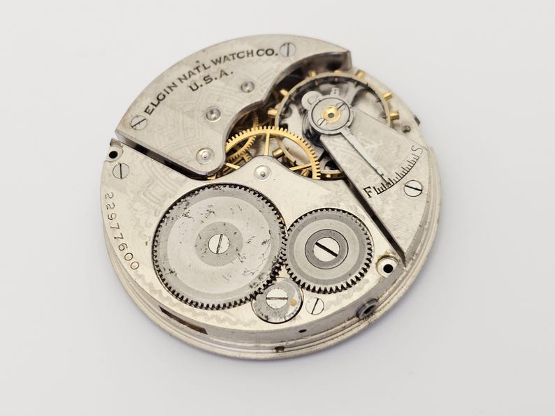 以下が含まれることがあります： Elgin National Watch Co. U.S.A. の時計ムーブメントのクローズアップ。 銀色のメカニズムは、複雑な歯車、ネジ、番号2297600を表示します。 このヴィンテージの時計ムーブメントは、時計の内部コンポーネントを明らかにしています。