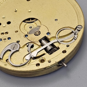 May include: Close-up of a gold-colored pocket watch mechanism with intricate gears and levers. The watch face is not visible.