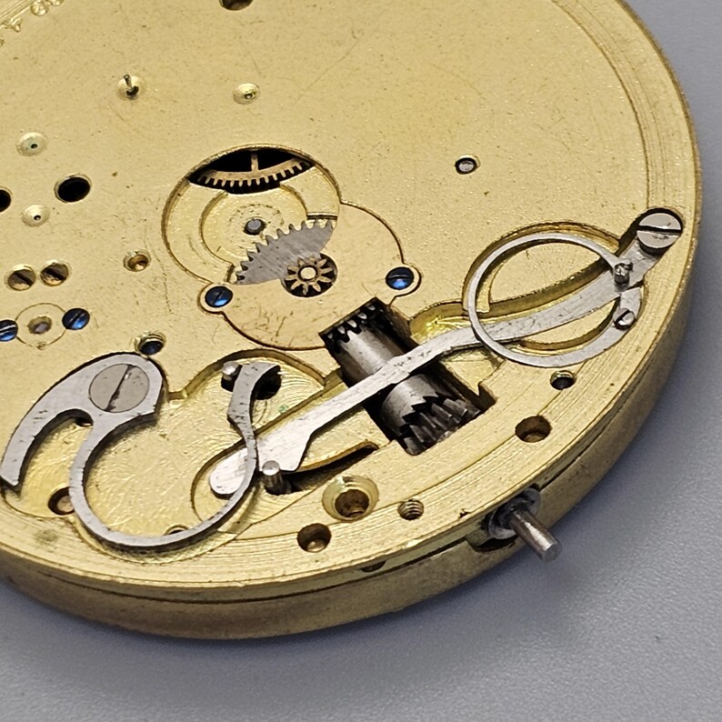 May include: Close-up of a gold-colored pocket watch mechanism with intricate gears and levers. The watch face is not visible.