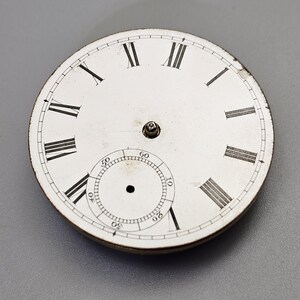 Puede incluir: Esfera de reloj blanca con marcadores de hora en n&uacute;meros romanos negros y marcadores de minutos negros alrededor del borde exterior. El centro de la esfera tiene un peque&ntilde;o c&iacute;rculo negro con un punto blanco en el centro.