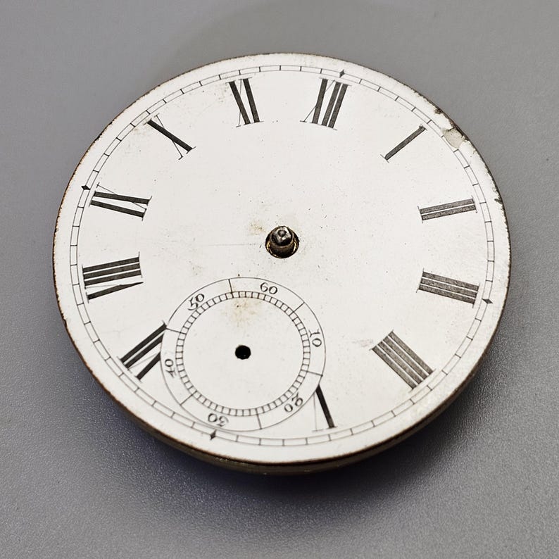 Puede incluir: Esfera de reloj blanca con marcadores de hora en n&uacute;meros romanos negros y marcadores de minutos negros alrededor del borde exterior. El centro de la esfera tiene un peque&ntilde;o c&iacute;rculo negro con un punto blanco en el centro.