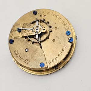 May include: Close-up of a vintage Elgin Natl. Watch Co. pocket watch movement. The gold-toned mechanism features intricate gears, blue screws, and the engraved text "Elgin Ill." and "1404669".
