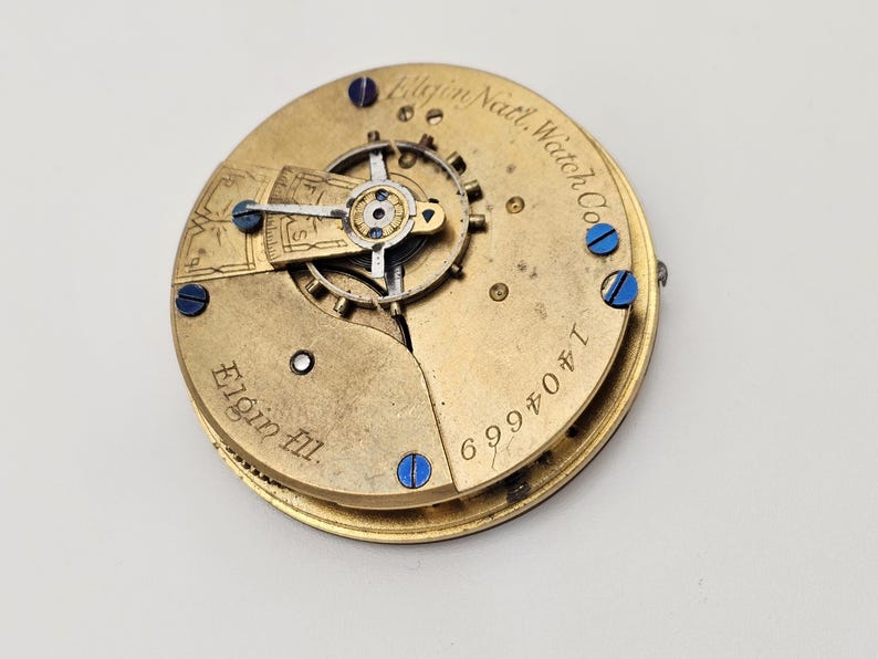 May include: Close-up of a vintage Elgin Natl. Watch Co. pocket watch movement. The gold-toned mechanism features intricate gears, blue screws, and the engraved text "Elgin Ill." and "1404669".