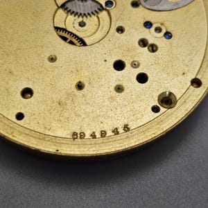 May include: Close-up of a gold-colored watch movement with the number 694945 engraved on the bottom.