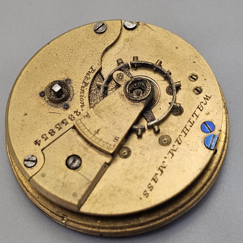 Puede incluir: Primer plano de un movimiento de reloj de bolsillo de color dorado con la inscripci&oacute;n "Waltham, Mass. E.T.C. Co. Patent, 18884" visible en la parte posterior.
