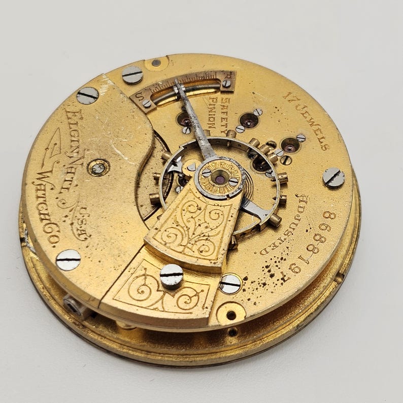 May include: Close-up of a vintage Elgin National Watch Co. pocket watch movement. The gold-toned mechanism displays intricate gears, a balance wheel, and the text "17 Jewels." The serial number 8688197 is visible. The watch is marked "Adjusted" and "Safety Pinion."