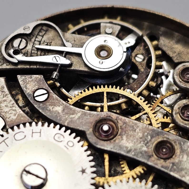 May include: Close-up of a vintage watch mechanism. The image showcases intricate gears, springs, and levers in shades of gold, silver, and bronze. The watch components are arranged in a complex, detailed pattern, highlighting the craftsmanship of the timepiece.