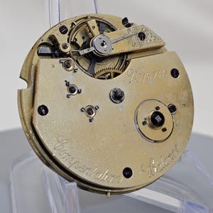 Może przedstawiać: Zbliżenie na mechanizm kieszonkowego zegarka w kolorze złotym z napisem "Compensation Balance" i numerem "1672740".