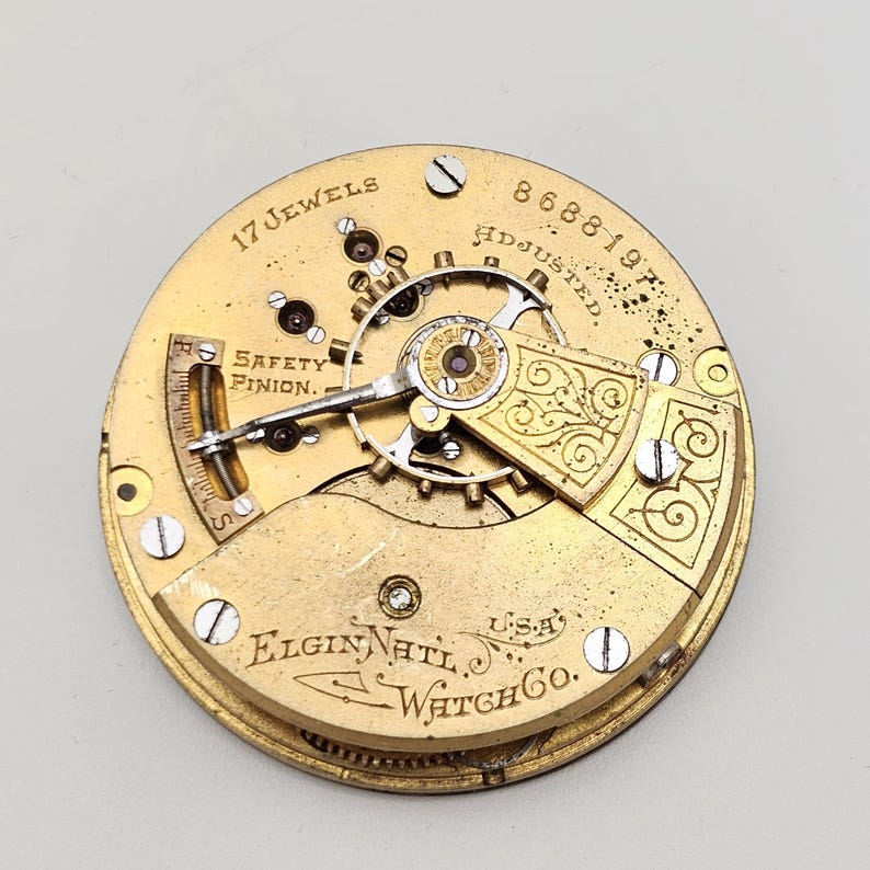 May include: Close-up of a vintage Elgin National Watch Co. movement. The gold-toned mechanism features intricate gears, the text "17 Jewels," and "Safety Pinion." The serial number "8688197" is visible. The watch movement is a piece of horological history.