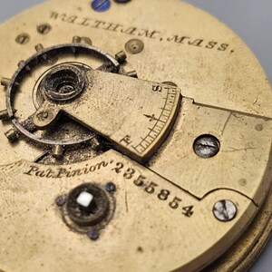 Puede incluir: Primer plano de un movimiento de reloj de color dorado con el texto "WALTHAM, MASS." y "Pat. Pinion' 2355854".