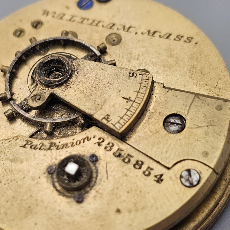 Puede incluir: Primer plano de un movimiento de reloj de color dorado con el texto "WALTHAM, MASS." y "Pat. Pinion' 2355854".