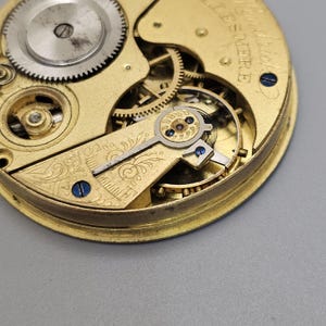 May include: Close-up of a gold-toned pocket watch movement with intricate gears and a decorative floral design. The watch is labeled "Swiss Made" and "15 Jewel."