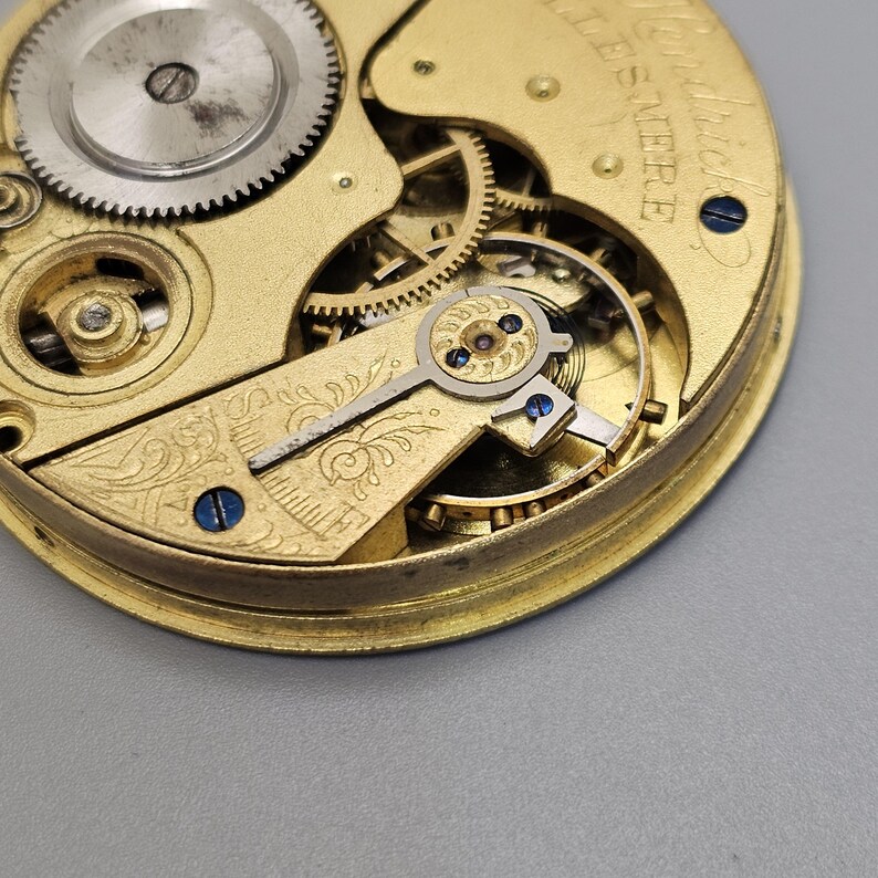May include: Close-up of a gold-toned pocket watch movement with intricate gears and a decorative floral design. The watch is labeled "Swiss Made" and "15 Jewel."