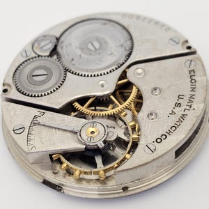 以下が含まれることがあります： ヴィンテージのElgin National Watch Co.のムーブメントのクローズアップ。銀色のメカニズムは、複雑な歯車、ネジ、テンプを備えています。「Elgin National Watch Co. U.S.A.」の文字が見えます。この時計のムーブメントは、20世紀初頭の時計製造の精巧な例です。