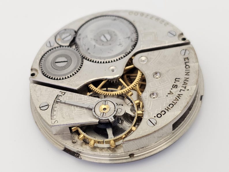以下が含まれることがあります： ヴィンテージのElgin National Watch Co.のムーブメントのクローズアップ。銀色のメカニズムは、複雑な歯車、ネジ、テンプを備えています。「Elgin National Watch Co. U.S.A.」の文字が見えます。この時計のムーブメントは、20世紀初頭の時計製造の精巧な例です。