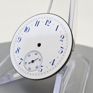 Puede incluir: Esfera de reloj de bolsillo blanca con marcadores de hora en n&uacute;meros romanos azules y agujas negras. La esfera del reloj tiene un subdial circular m&aacute;s peque&ntilde;o con agujas negras y marcas blancas.