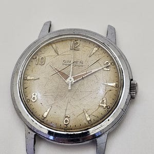 May include: Vintage silver-toned watch with a cream-colored face. The watch face features black numerals and hands, with the brand name "GREEN PROVISION" printed at the top. The watch has a scratched appearance, suggesting age.