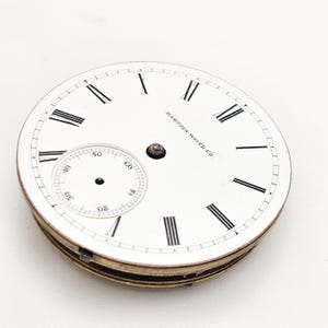 Puede incluir: Una esfera de reloj de bolsillo vintage con esfera blanca y n&uacute;meros romanos negros. La esfera incluye una esfera secundaria m&aacute;s peque&ntilde;a y el texto "HAMPDEN WATCH CO." El reloj tiene un borde met&aacute;lico.
