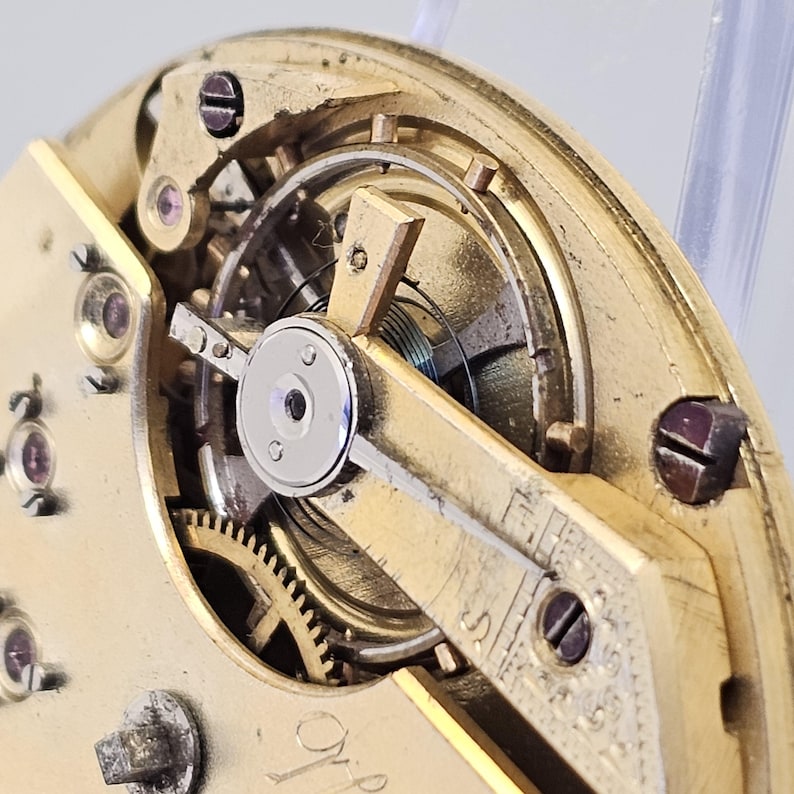 Może przedstawiać: Zbliżenie na złoty mechanizm zegarka z misternymi kołami zębatymi i mechanizmem naciągu. Mechanizm jest widoczny przez przezroczystą szklaną pokrywę.