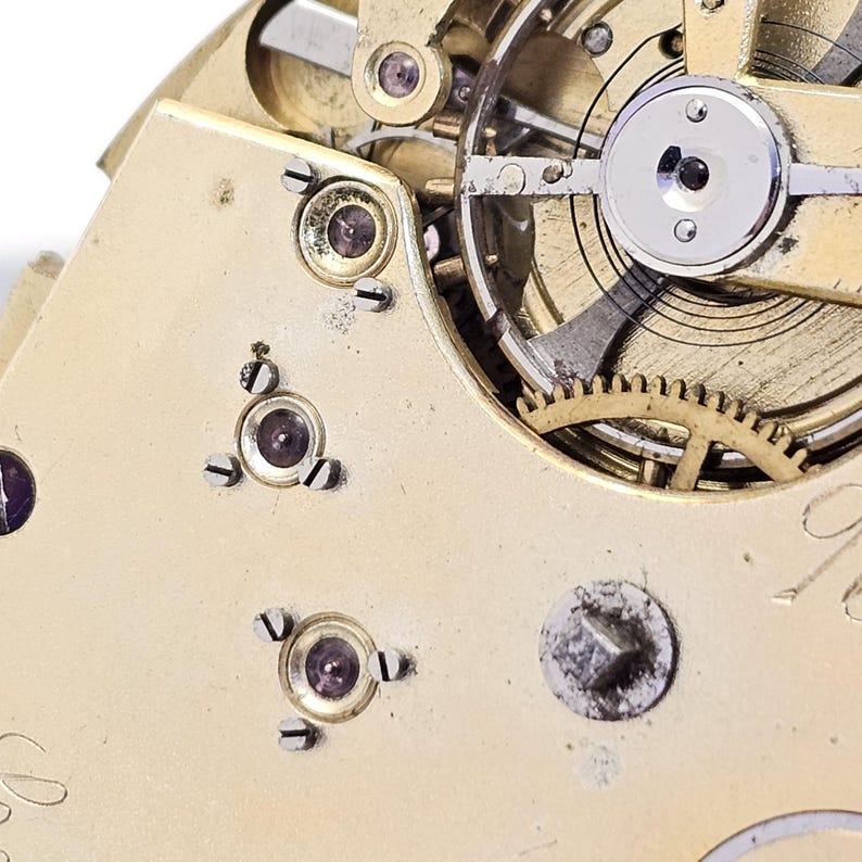 Może przedstawiać: Zbliżenie na mechanizm kieszonkowego zegarka w kolorze złotym z widocznymi kołami zębatymi i śrubami. Tarcza zegarka nie jest widoczna na zdjęciu.