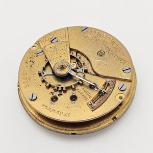 May include: A vintage watch movement with intricate gears and components. The gold-toned mechanism features the text "17 Jewels" and "Elgin Natl. Watch Co." along with other markings. The watch movement is round and has a complex design.