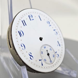 Puede incluir: Una esfera de reloj de bolsillo blanca con marcadores de hora en n&uacute;meros romanos azules y un subdial blanco m&aacute;s peque&ntilde;o con manecillas negras. La esfera del reloj tiene un borde dorado con un patr&oacute;n de puntos.
