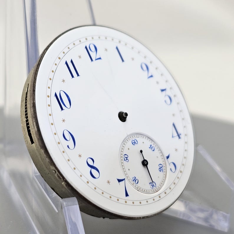 Puede incluir: Una esfera de reloj de bolsillo blanca con marcadores de hora en n&uacute;meros romanos azules y un subdial blanco m&aacute;s peque&ntilde;o con manecillas negras. La esfera del reloj tiene un borde dorado con un patr&oacute;n de puntos.