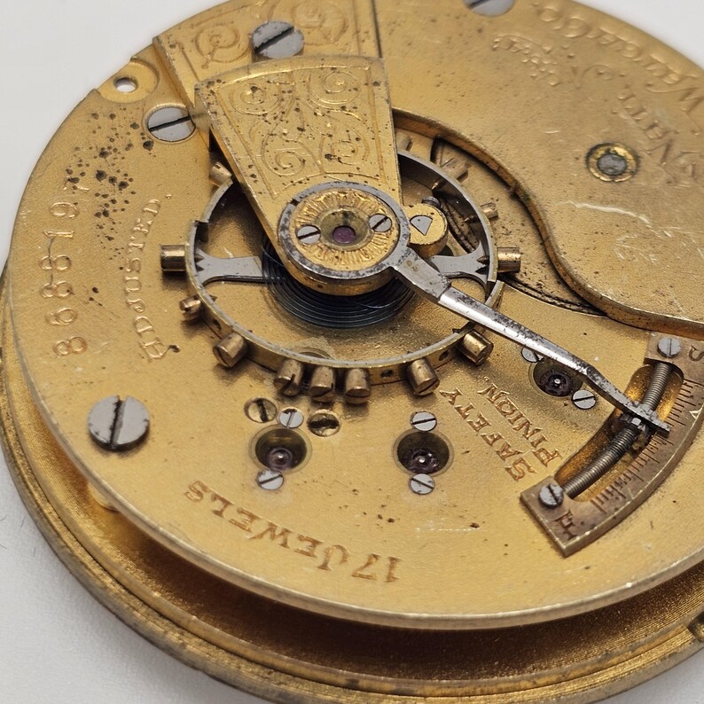May include: Close-up of a vintage watch movement with intricate gold-toned gears, screws, and a balance wheel. The watch mechanism features the text "17 Jewels" and "Safety Pinion". The overall design is detailed and shows signs of age.