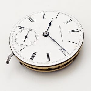 May include: A vintage pocket watch face with a white dial, black Roman numerals, and a smaller seconds dial. The watch hands are black, and the outer casing is gold-toned. The text "10 NATIONAL TIME PIECE" is visible.