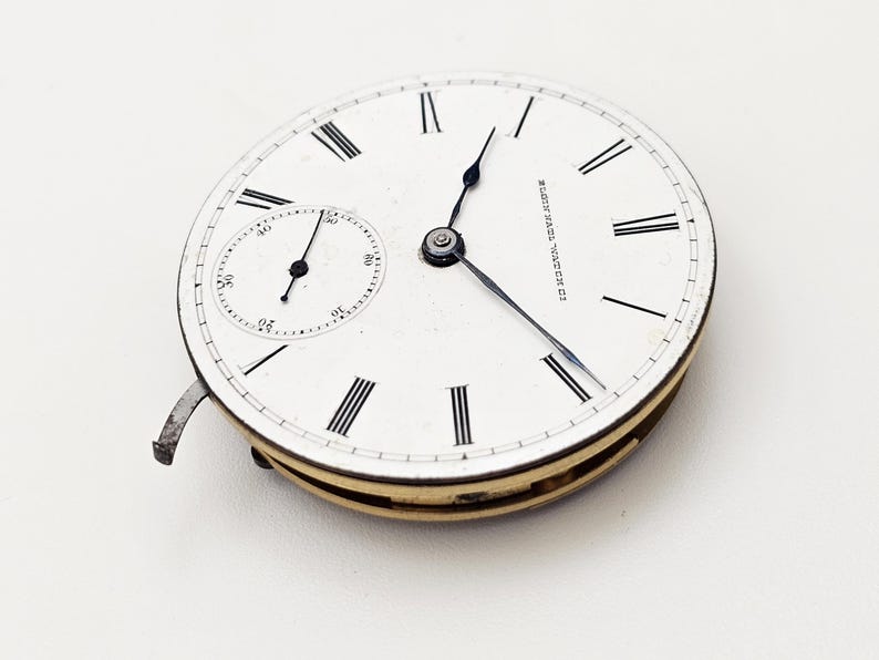 May include: A vintage pocket watch face with a white dial, black Roman numerals, and a smaller seconds dial. The watch hands are black, and the outer casing is gold-toned. The text "10 NATIONAL TIME PIECE" is visible.