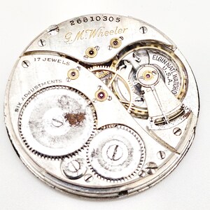 May include: Close-up of a vintage watch movement with intricate gears and components. The watch mechanism is silver-toned, with the text "G.M. Wheeler" and "Elgin Natl Watch Co U.S.A." visible. Features "17 Jewels" and "Six Adjustments".