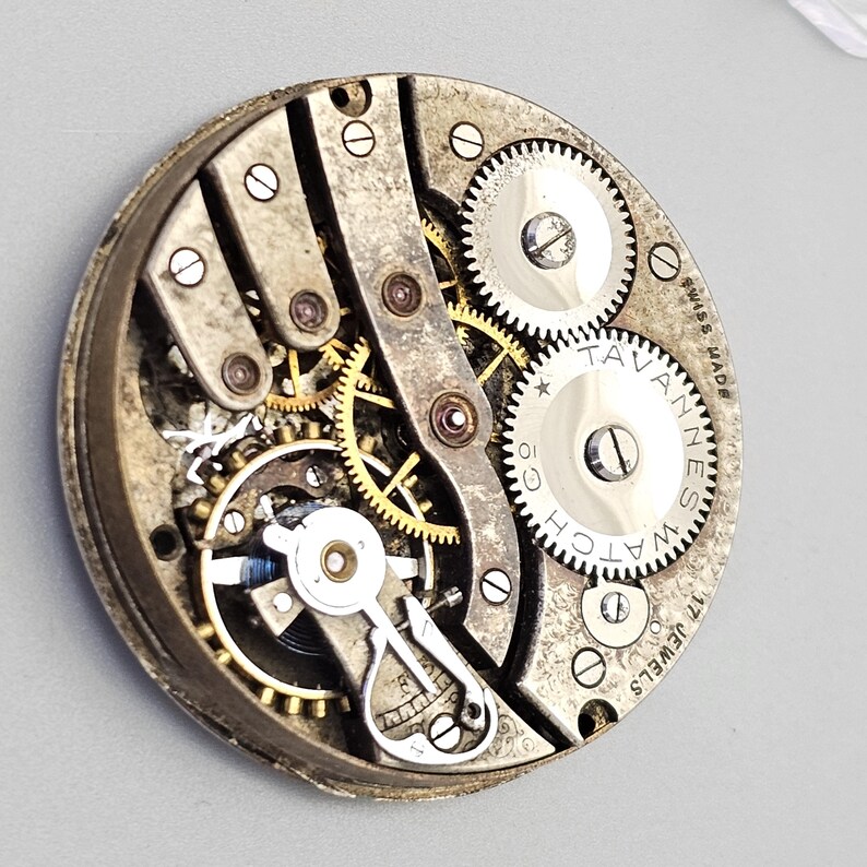 May include: Close-up of a vintage watch movement, showcasing intricate gears and mechanisms. The watch components are silver and gold, with the words "Tavannes Watch" and "Swiss Made" visible. The watch has 17 jewels.