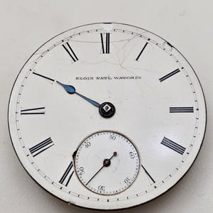 May include: Antique Elgin National Watch Co. pocket watch face. The white face features black Roman numerals, a smaller seconds dial, and blue hands. The watch face has visible cracks.