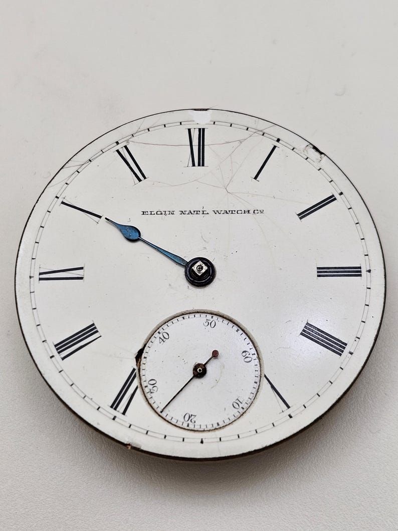 May include: Antique Elgin National Watch Co. pocket watch face. The white face features black Roman numerals, a smaller seconds dial, and blue hands. The watch face has visible cracks.