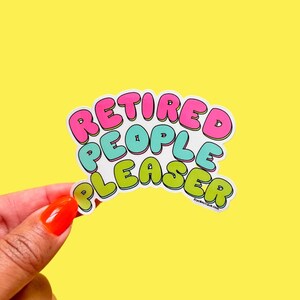 May include: A sticker with the text "Retired People Pleaser" in a rainbow of colours. The sticker is held in a hand.