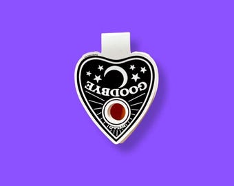 May include: A heart-shaped, black and white Ouija board planchette with the word "GOODBYE" in white, a crescent moon, and stars. It has a red, circular viewing window and a white tab at the top.