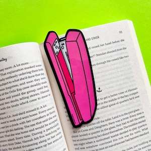 May include: A pink plastic stapler bookmark with a black outline. The bookmark is placed on an open book with text about a bar and a game of quarters.