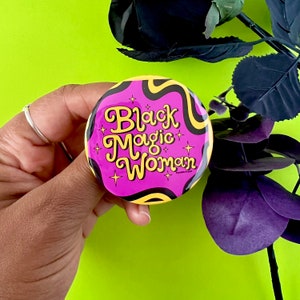 May include: A pink and yellow button with the text "Black Magic Woman" in a retro font. The button has a swirl pattern.