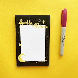 Op de afbeelding: Zwarte notitieblok met gouden opschrift "Spells & Potions", een halve maan en sterren. Het notitieblok ligt op een gele ondergrond met een roze en grijze Sharpie-marker.