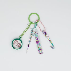 May include: A green keychain with a variety of colorful charms, including a small round mirror, a pink safety pin, and three colorful crochet hooks.