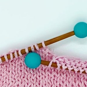 May include: Close-up of pink knitting with two turquoise knitting needles. The needles have turquoise plastic tips.