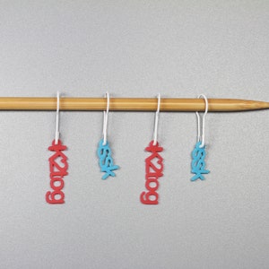 May include: Four stitch markers hanging on a wooden knitting needle. Two markers are red with the text "k2tog" and two are blue with the text "ssk".