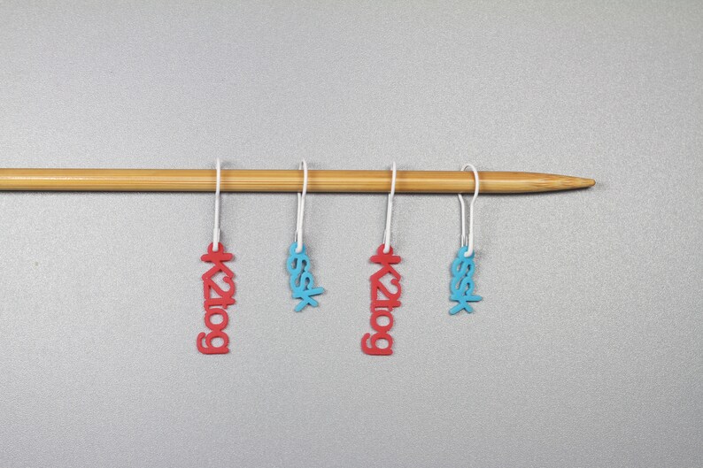 May include: Four stitch markers hanging on a wooden knitting needle. Two markers are red with the text "k2tog" and two are blue with the text "ssk".