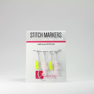 May include: A set of four stitch markers with pink and yellow accents. The markers are labelled "K2tog" and "azadas knitting accessories".