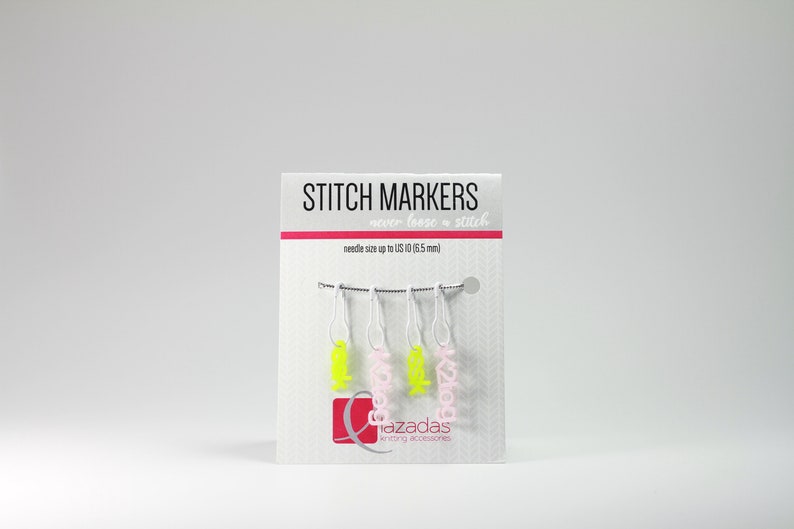 May include: A set of four stitch markers with pink and yellow accents. The markers are labelled "K2tog" and "azadas knitting accessories".